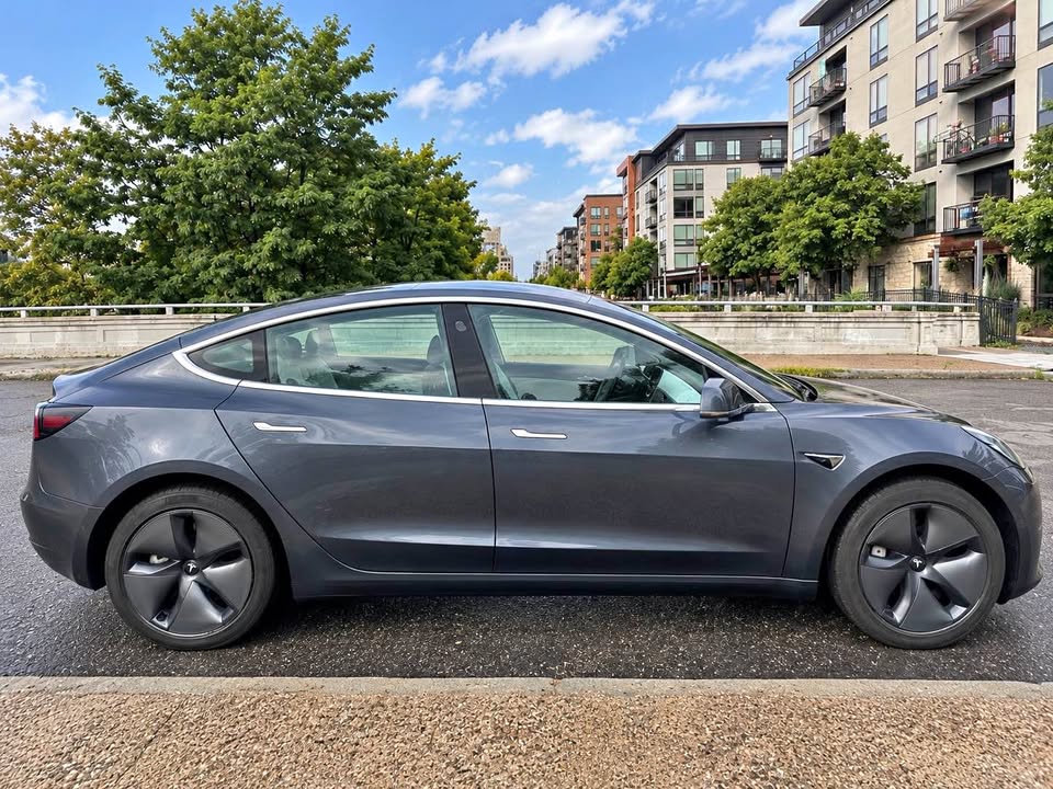 Tesla Model 3 in dark gray metallic paint, side profile on a city street with trees and apartments in the background